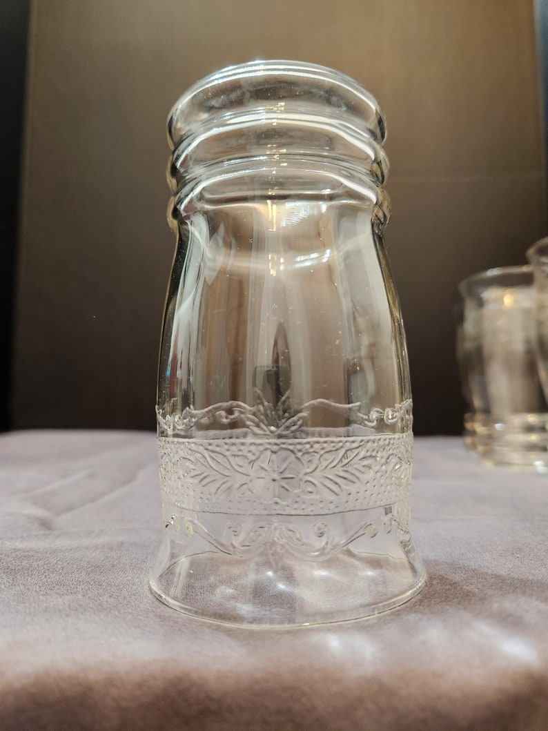 Puede incluir: Un vaso de beber de vidrio transparente con un patr&oacute;n floral decorativo alrededor del medio. El vaso tiene una base ancha y una parte superior ligeramente acampanada.