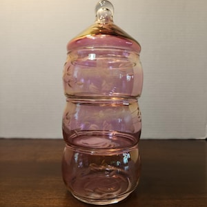 May include: A three-tiered, iridescent pink glass jar with a lid. The jar has a decorative floral pattern etched into the glass. The lid has a clear glass knob. The jar is likely used for storing small items.