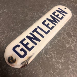Puede incluir: Un letrero rectangular vintage con la palabra "GENTLEMEN" en letras azul marino sobre un fondo blanco. El letrero tiene bordes redondeados y un pequeño agujero en un extremo para colgar. El letrero tiene un aspecto envejecido.