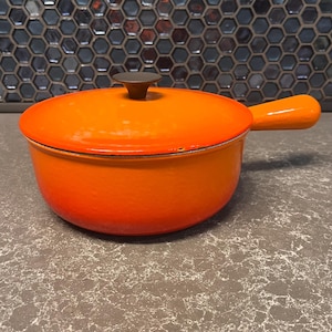 Pode incluir: Panela de ferro fundido esmaltado laranja brilhante com tampa e alça. A panela tem um botão marrom escuro na tampa e uma alça laranja combinando. A panela está em uma bancada cinza. Mede cerca de 25 cm de diâmetro.