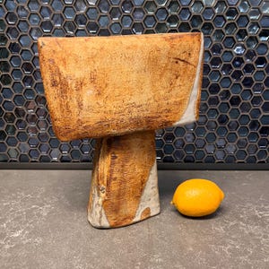 May include: A handmade ceramic vase with a rectangular top and a pedestal base. The vase is tan with white accents and a rough texture. A bright yellow lemon sits beside the vase. The vase is approximately 20 cm tall.