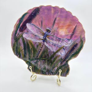 May include: A painted scallop shell featuring a dragonfly with translucent wings resting among green reeds against a purple and orange sunset background. The shell has a gold-painted scalloped edge and is displayed on a gold metal wire stand.