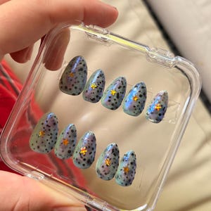 May include: A set of oval-shaped false nails in a clear plastic case. The nails have a blue-green iridescent base with black speckles and gold star accents. The nails are designed for cosmetic use.