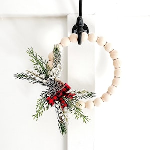 Puede incluir: Una corona decorativa de cuentas de madera con un arreglo festivo de vegetaci&oacute;n artificial, pi&ntilde;as y un lazo a cuadros rojo y negro. Cuelga de un gancho de metal negro sobre un fondo blanco.