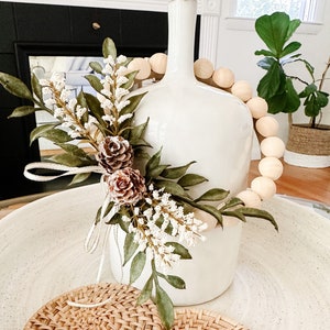 K&ouml;nnte beinhalten: Eine wei&szlig;e Keramikvase mit einem dekorativen Blumenarrangement und einer Holzperlen-Girlande. Zwei gewebte Rattan-Untersetzer liegen in einer gesprenkelten wei&szlig;en Schale. Das Arrangement umfasst gr&uuml;ne Bl&auml;tter, wei&szlig;e Beeren und braune Tannenzapfen.