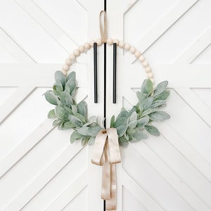 May include: A wooden bead wreath with a green leafy garland and a gold ribbon hanging on a white door with a geometric pattern.
