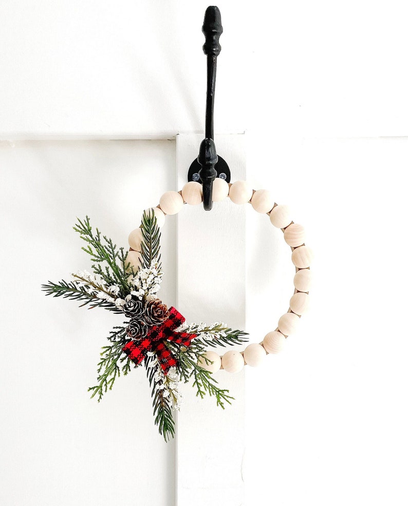 Puede incluir: Una corona decorativa de cuentas de madera con un arreglo festivo de vegetaci&oacute;n artificial, pi&ntilde;as y un lazo a cuadros rojo y negro. Cuelga de un gancho de metal negro sobre un fondo blanco.