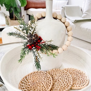 Puede incluir: Un jarr&oacute;n de cer&aacute;mica blanco adornado con una guirnalda de cuentas de madera y un arreglo festivo de pino, bayas y pi&ntilde;as. Tres posavasos de rat&aacute;n tejido descansan en un cuenco blanco, creando una exhibici&oacute;n acogedora y de temporada.