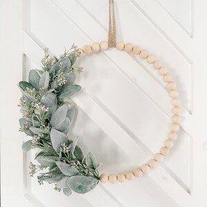 May include: A wooden bead wreath with a green and white leafy garland attached. The wreath is hanging on a white door with a diagonal pattern.