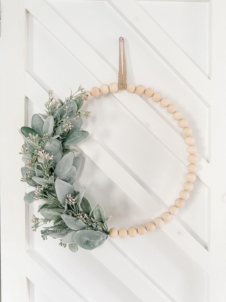 May include: A wooden bead wreath with a green and white leafy garland attached. The wreath is hanging on a white door with a diagonal pattern.