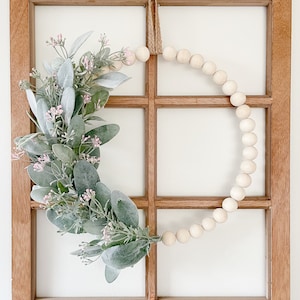 May include: A wooden window frame with a wreath made of green leaves and pink flowers. The wreath is decorated with a string of white wooden beads.