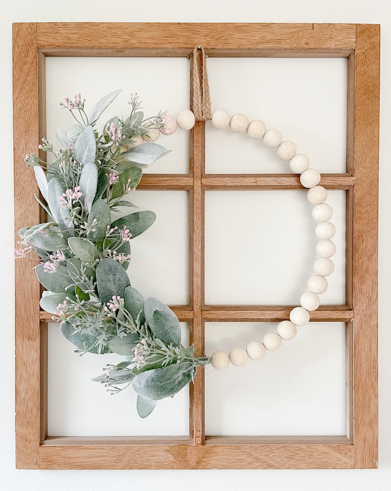 May include: A wooden window frame with a wreath made of green leaves and pink flowers. The wreath is decorated with a string of white wooden beads.