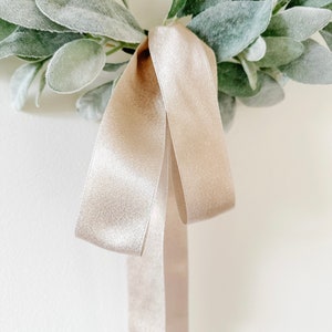May include: A simple, rustic wreath made of green leaves with a beige satin ribbon tied in a bow.