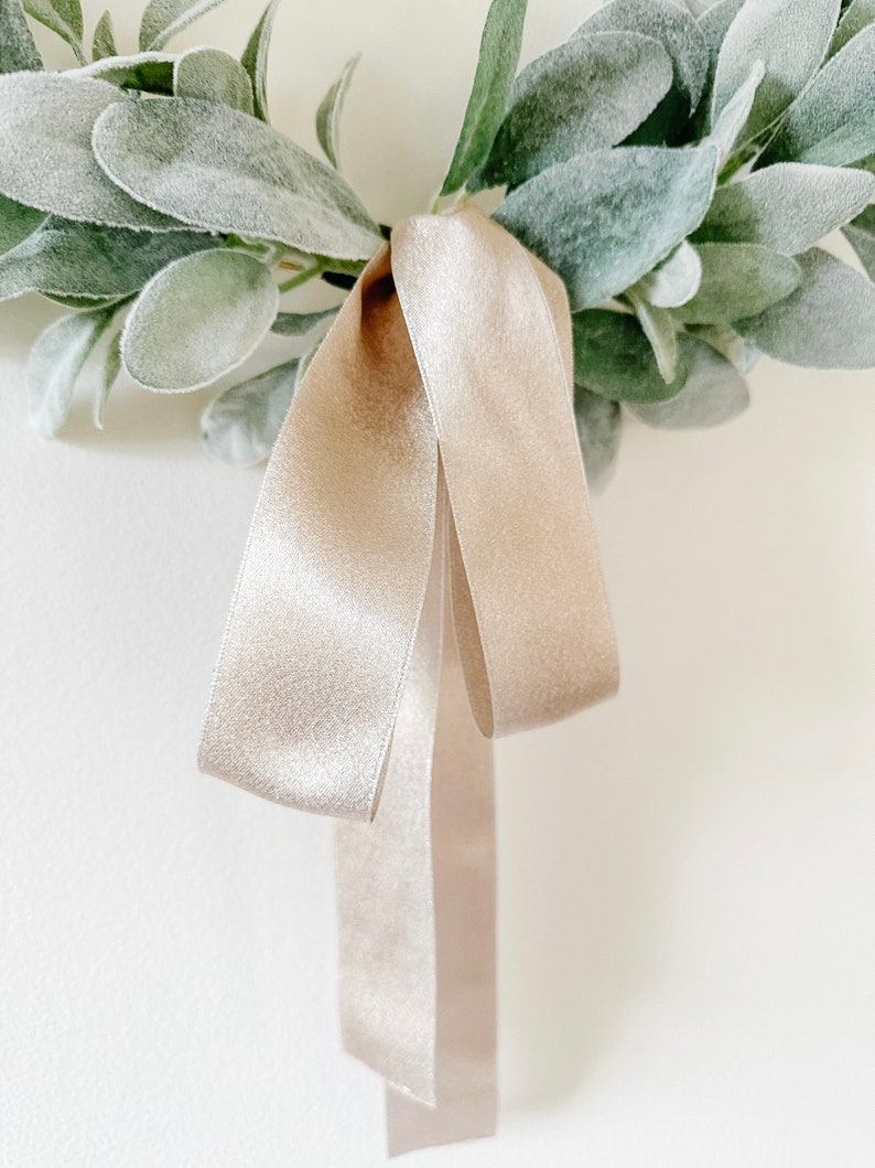 May include: A simple, rustic wreath made of green leaves with a beige satin ribbon tied in a bow.