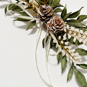 K&ouml;nnte beinhalten: Nahaufnahme eines dekorativen Blumenarrangements mit zwei Tannenzapfen, gr&uuml;nen Bl&auml;ttern und kleinen wei&szlig;en Bl&uuml;ten. Ein wei&szlig;es Band mit goldenem Besatz ist um das Arrangement gebunden. Das Arrangement befindet sich vor einem wei&szlig;en Hintergrund.