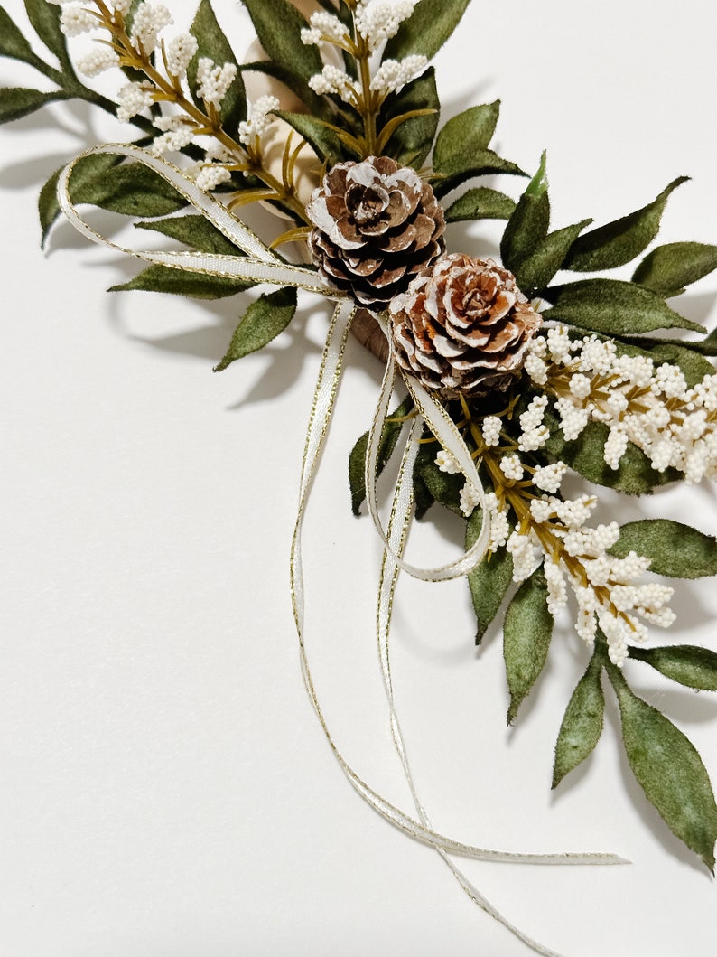 K&ouml;nnte beinhalten: Nahaufnahme eines dekorativen Blumenarrangements mit zwei Tannenzapfen, gr&uuml;nen Bl&auml;ttern und kleinen wei&szlig;en Bl&uuml;ten. Ein wei&szlig;es Band mit goldenem Besatz ist um das Arrangement gebunden. Das Arrangement befindet sich vor einem wei&szlig;en Hintergrund.