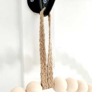 May include: A wooden bead garland with a jute rope hanger. The garland is hanging from a black hook on a white wall.