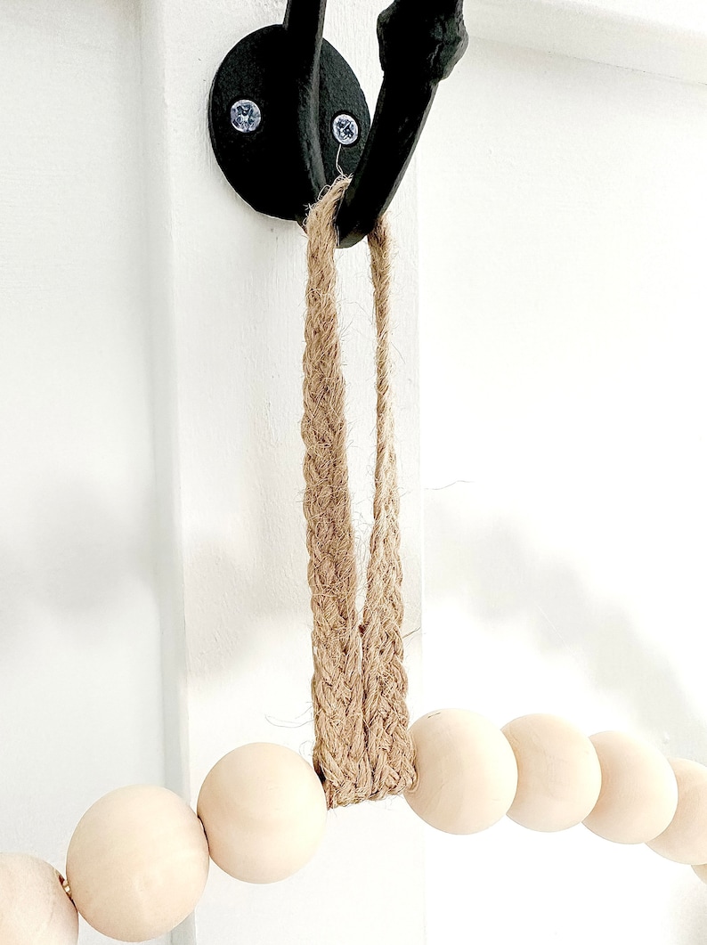 May include: A wooden bead garland with a jute rope hanger. The garland is hanging from a black hook on a white wall.