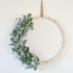 May include: A white beaded wreath with a green and white floral arrangement. The wreath is hanging from a brown twine loop.