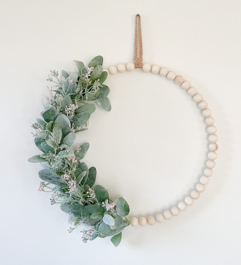 May include: A white beaded wreath with a green and white floral arrangement. The wreath is hanging from a brown twine loop.