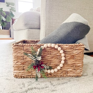 Puede incluir: Una cesta rectangular tejida con una corona decorativa de cuentas de madera y un peque&ntilde;o arreglo de pi&ntilde;as y vegetaci&oacute;n. La cesta contiene una manta gris y blanca. La cesta est&aacute; sobre una alfombra gris clara.