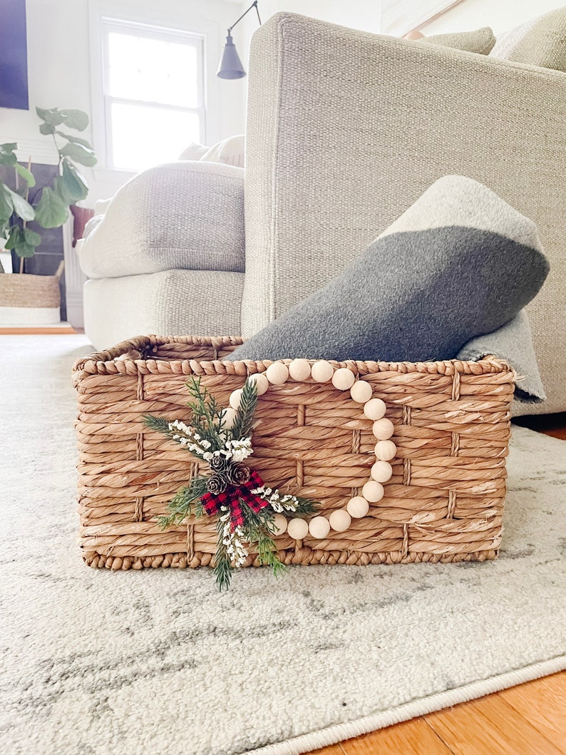 Puede incluir: Una cesta rectangular tejida con una corona decorativa de cuentas de madera y un peque&ntilde;o arreglo de pi&ntilde;as y vegetaci&oacute;n. La cesta contiene una manta gris y blanca. La cesta est&aacute; sobre una alfombra gris clara.