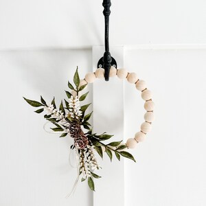 K&ouml;nnte beinhalten: Ein dekorativer Holzkranz mit floralen und Tannenzapfen-Akzenten. Der Kranz ist mit gr&uuml;nen Bl&auml;ttern, wei&szlig;en Bl&uuml;ten und braunen Tannenzapfen verziert. Er h&auml;ngt an einem schwarzen Metallhaken vor einem wei&szlig;en Hintergrund.