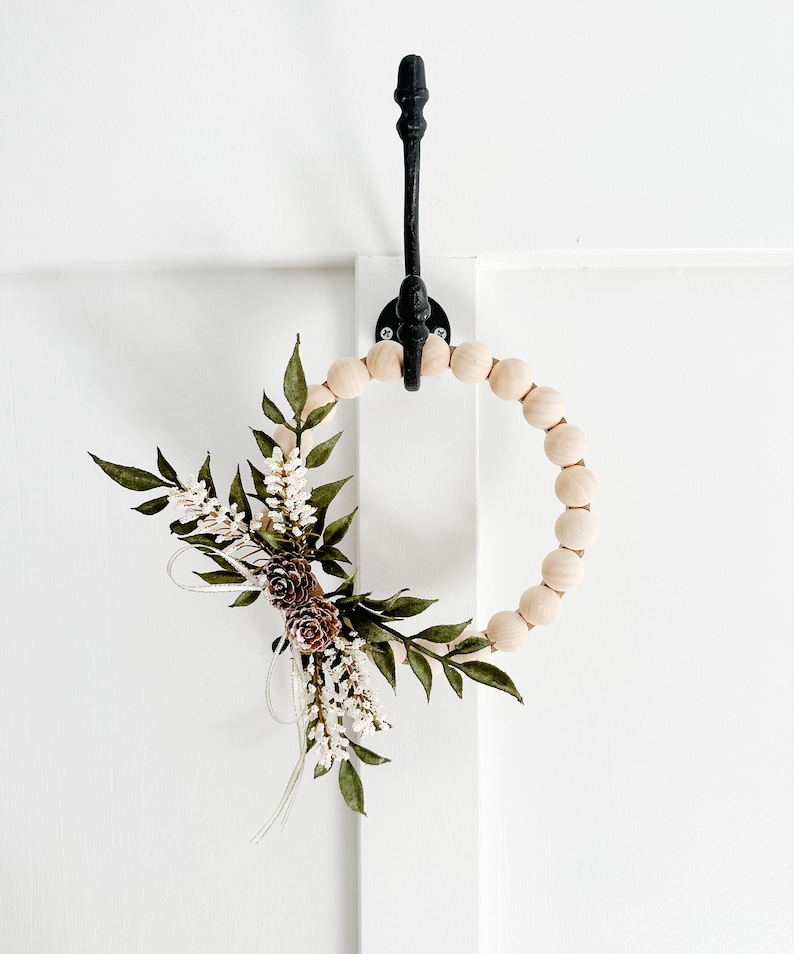 K&ouml;nnte beinhalten: Ein dekorativer Holzkranz mit floralen und Tannenzapfen-Akzenten. Der Kranz ist mit gr&uuml;nen Bl&auml;ttern, wei&szlig;en Bl&uuml;ten und braunen Tannenzapfen verziert. Er h&auml;ngt an einem schwarzen Metallhaken vor einem wei&szlig;en Hintergrund.