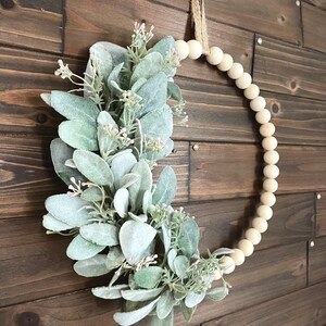 May include: A light green and white faux lamb's ear wreath with a natural twine hanger. The wreath is made of wooden beads and is hanging on a brown wooden wall.