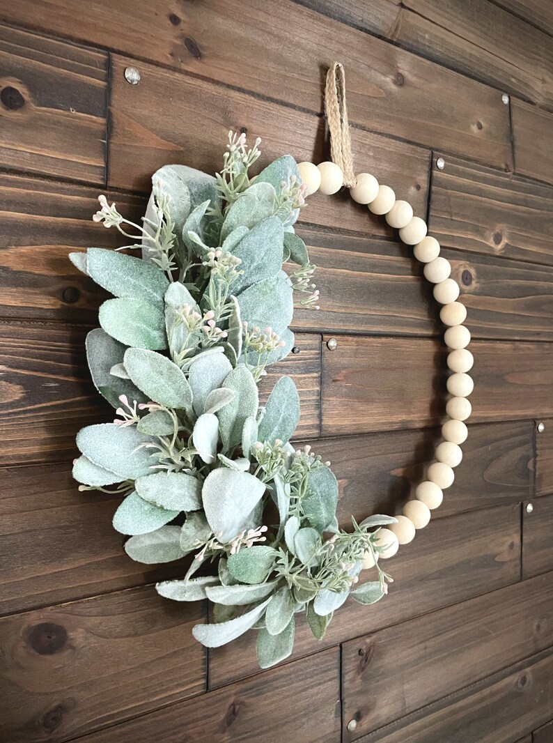 May include: A light green and white faux lamb's ear wreath with a natural twine hanger. The wreath is made of wooden beads and is hanging on a brown wooden wall.