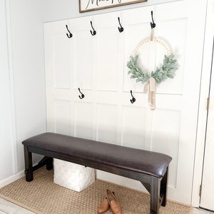 May include: A brown leather-topped bench with a white wicker basket underneath. The bench is in front of a white wall with a wooden sign that says "MacNeil" and black hooks. A wreath with greenery and a beige ribbon hangs on the wall.