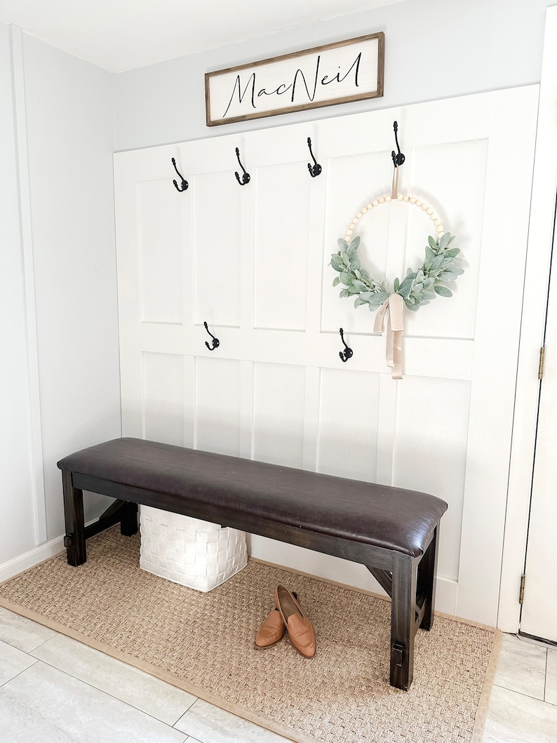 May include: A brown leather-topped bench with a white wicker basket underneath. The bench is in front of a white wall with a wooden sign that says "MacNeil" and black hooks. A wreath with greenery and a beige ribbon hangs on the wall.