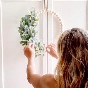 May include: A person is hanging a white beaded wreath with a green and white floral arrangement on a white cabinet door.