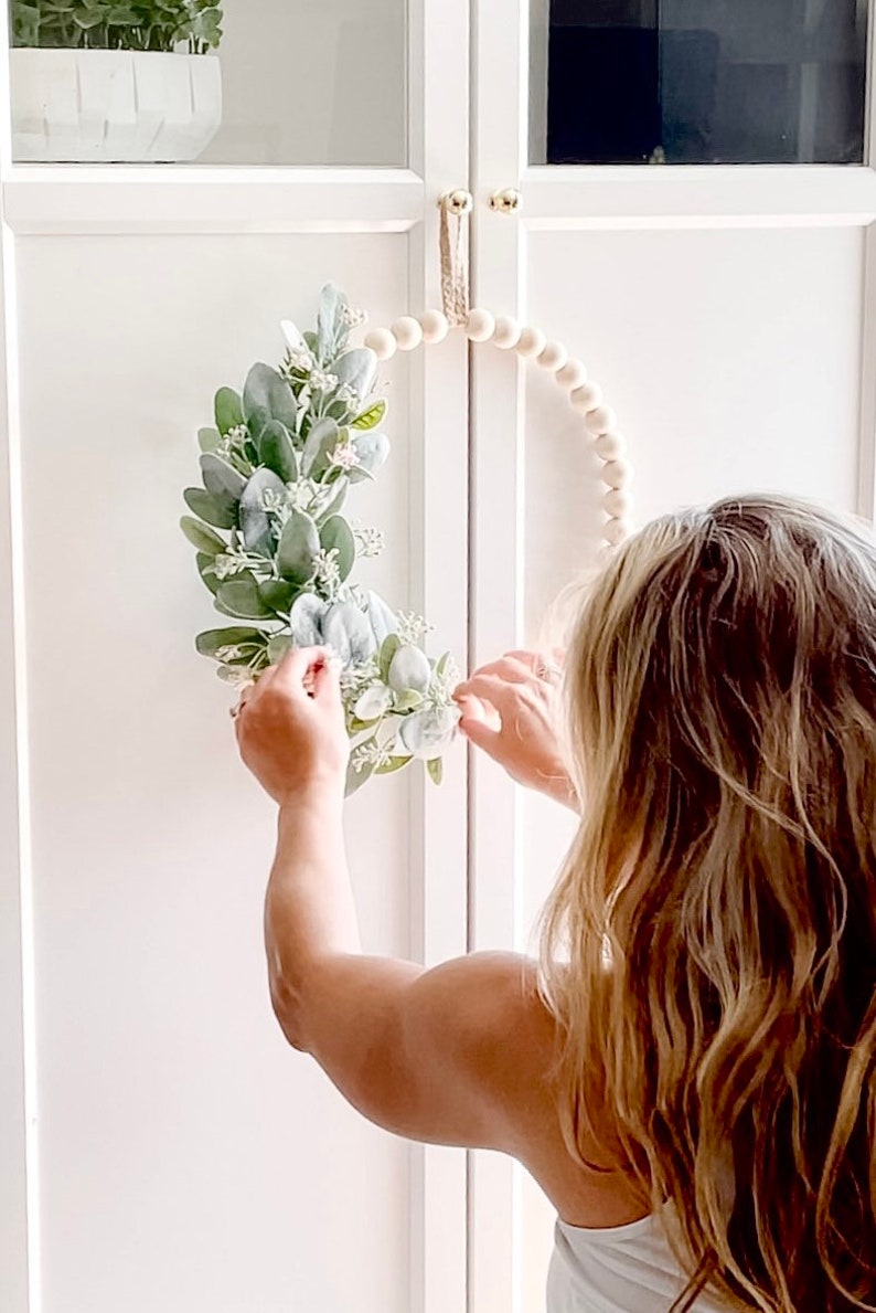 May include: A person is hanging a white beaded wreath with a green and white floral arrangement on a white cabinet door.