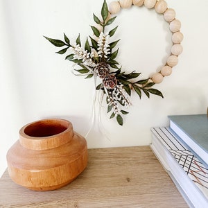 K&ouml;nnte beinhalten: Ein kreisf&ouml;rmiger Kranz aus Holzperlen, verziert mit gr&uuml;nen Bl&auml;ttern, wei&szlig;en Bl&uuml;ten und Tannenzapfen. Er h&auml;ngt an einer wei&szlig;en Wand. Eine Holzvase und B&uuml;cher stehen auf einer Holzoberfl&auml;che.