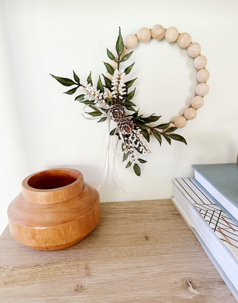 K&ouml;nnte beinhalten: Ein kreisf&ouml;rmiger Kranz aus Holzperlen, verziert mit gr&uuml;nen Bl&auml;ttern, wei&szlig;en Bl&uuml;ten und Tannenzapfen. Er h&auml;ngt an einer wei&szlig;en Wand. Eine Holzvase und B&uuml;cher stehen auf einer Holzoberfl&auml;che.