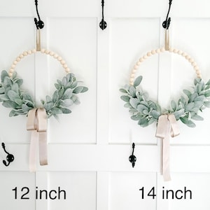 May include: Two wreaths made of wooden beads and greenery, with a beige ribbon hanging down. The wreath on the left is 12 inches in diameter, and the wreath on the right is 14 inches in diameter.