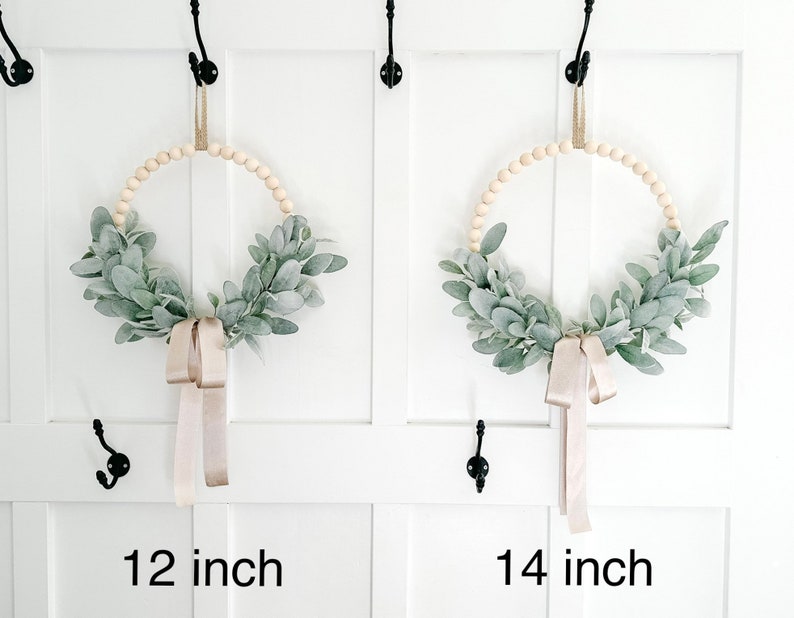 May include: Two wreaths made of wooden beads and greenery, with a beige ribbon hanging down. The wreath on the left is 12 inches in diameter, and the wreath on the right is 14 inches in diameter.