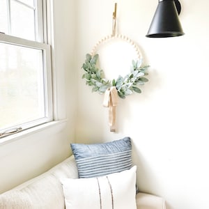 May include: A white couch with two pillows, one blue and white striped and one white with brown stripes. A wooden bead wreath with greenery hangs on the wall above the couch.