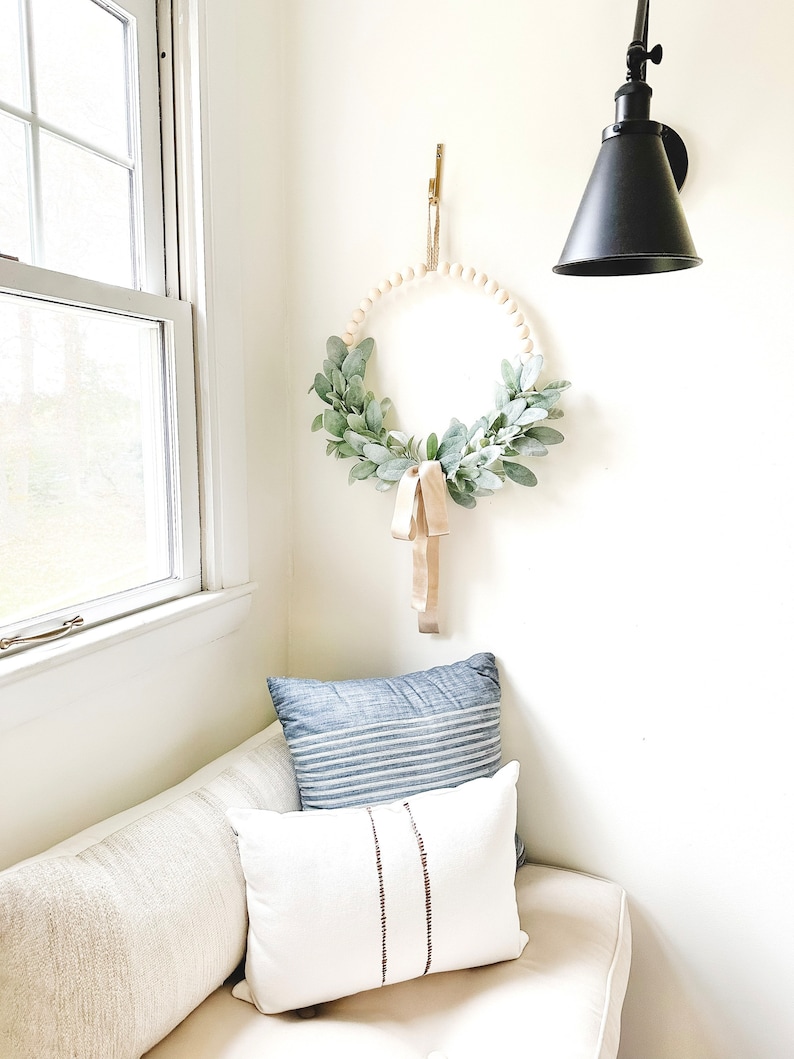 May include: A white couch with two pillows, one blue and white striped and one white with brown stripes. A wooden bead wreath with greenery hangs on the wall above the couch.
