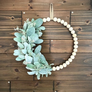 May include: A wreath made of white wooden beads with a green leafy garland attached to it. The wreath is hanging on a brown wooden wall.