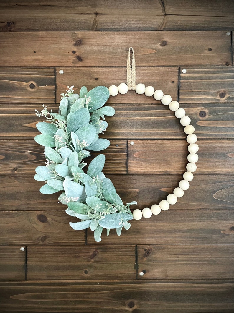 May include: A wreath made of white wooden beads with a green leafy garland attached to it. The wreath is hanging on a brown wooden wall.