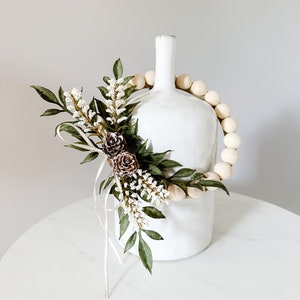 K&ouml;nnte beinhalten: Eine wei&szlig;e Keramikvase mit schmalem Hals, verziert mit einer Girlande aus Holzperlen. Die Vase ist mit k&uuml;nstlichem Gr&uuml;n, wei&szlig;en Bl&uuml;ten und kleinen Tannenzapfen geschm&uuml;ckt. Die Anordnung steht auf einer wei&szlig;en Oberfl&auml;che.