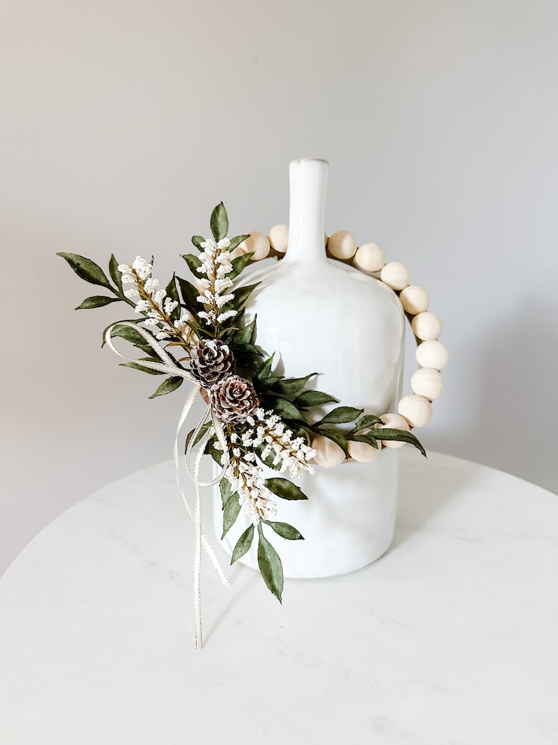 K&ouml;nnte beinhalten: Eine wei&szlig;e Keramikvase mit schmalem Hals, verziert mit einer Girlande aus Holzperlen. Die Vase ist mit k&uuml;nstlichem Gr&uuml;n, wei&szlig;en Bl&uuml;ten und kleinen Tannenzapfen geschm&uuml;ckt. Die Anordnung steht auf einer wei&szlig;en Oberfl&auml;che.