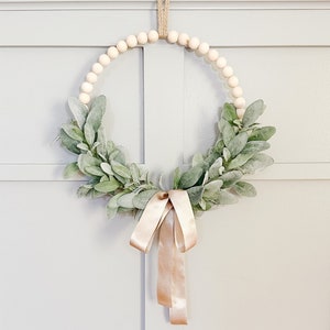 May include: A wooden bead wreath decorated with a light green leafy garland and a blush pink ribbon.