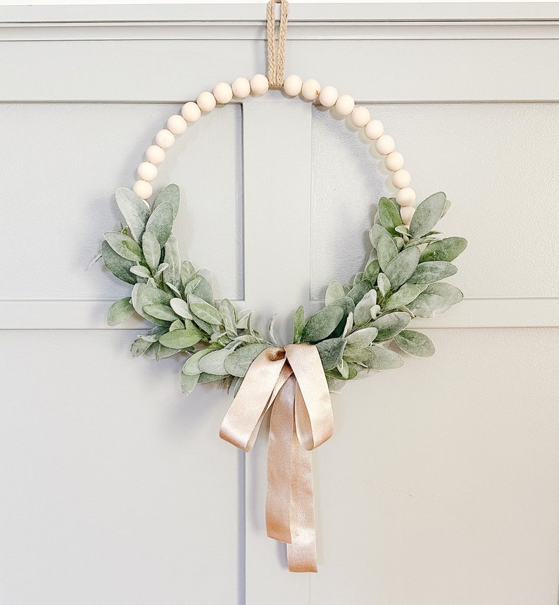 May include: A wooden bead wreath decorated with a light green leafy garland and a blush pink ribbon.