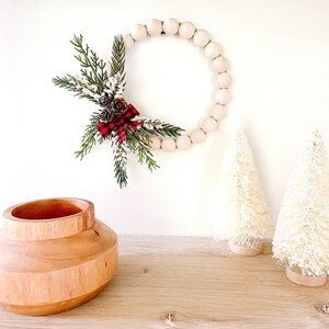Puede incluir: Una corona de cuentas de madera con pi&ntilde;as y un lazo a cuadros rojo y negro cuelga de una pared blanca. Dos peque&ntilde;os &aacute;rboles de Navidad blancos de cepillo para botellas y un jarr&oacute;n de madera se encuentran sobre una superficie de madera.