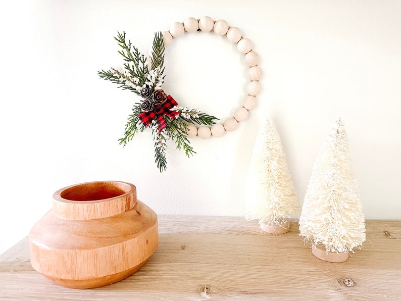 Puede incluir: Una corona de cuentas de madera con pi&ntilde;as y un lazo a cuadros rojo y negro cuelga de una pared blanca. Dos peque&ntilde;os &aacute;rboles de Navidad blancos de cepillo para botellas y un jarr&oacute;n de madera se encuentran sobre una superficie de madera.