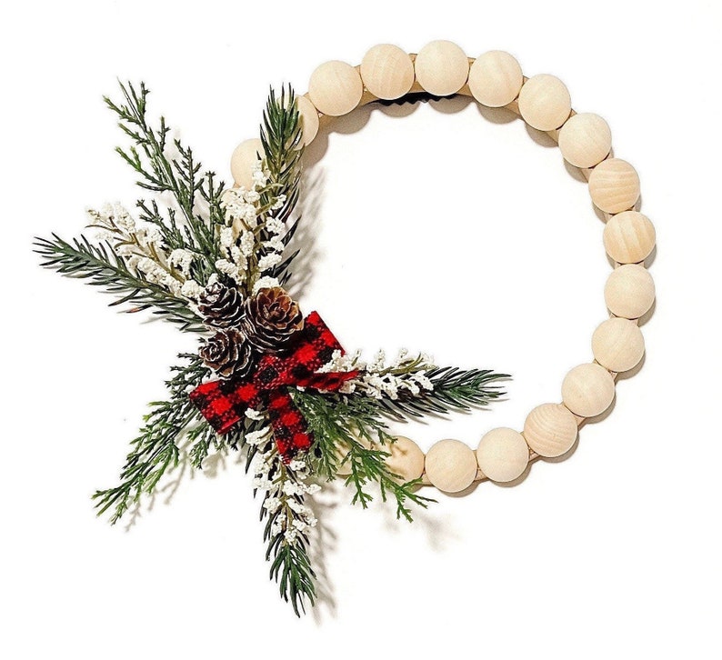 Puede incluir: Una corona circular de cuentas de madera, adornada con elementos festivos. Incluye ramas de pino verdes, acentos florales blancos y pi&ntilde;as marrones. Un lazo escoc&eacute;s rojo y negro proporciona contraste. Ideal para la decoraci&oacute;n de temporada.