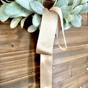 May include: A light beige ribbon with a shimmery texture hangs from a green wreath. The wreath is made of faux greenery and is hanging on a wooden wall.